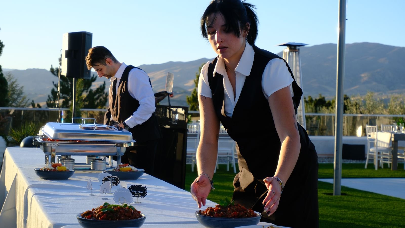 Bora staff laying out dishes on a buffet table outdoors, mountains behind